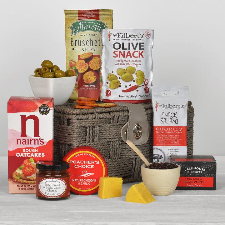 A curated gourmet snack and cheese hamper arranged in a woven basket on a light gray background. The basket is filled with packaged snacks including Nairn's Rough Oatcakes, Maretti Bruschetta Chips, Mr Filbert's Olive Snack, Mr Filbert's Snack Salami, Poacher's Choice Cheddar & Garlic, and Farmhouse Biscuits Black Pepper, alongside loose cheese wedges, a bowl of olives, and a small bowl of chutney.