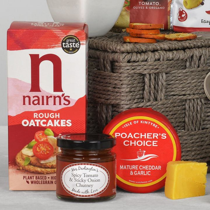 A styled food gift arrangement featuring a box of Nairn's Rough Oatcakes, a jar of Mrs Darlington's Spicy Tomato & Sticky Onion Chutney, a round Poacher's Choice Mature Cheddar & Garlic, and a wedge of yellow cheese, set against a woven basket and picnic-style background.