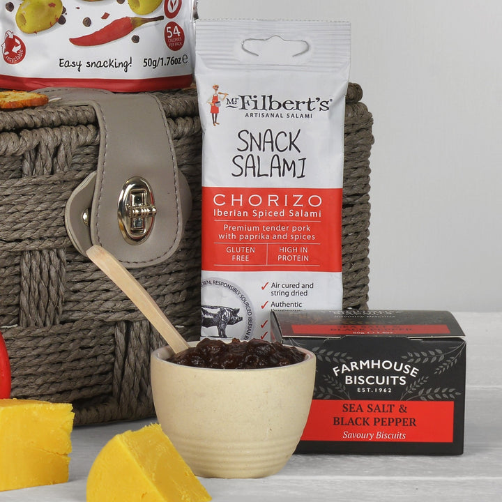 A curated snack and salami gift arrangement featuring a bag of Mr. Filbert's Chorizo Snack Salami, a bowl of chutney with a wooden spoon, a box of Farmhouse Biscuits Sea Salt & Black Pepper, and a woven grey basket