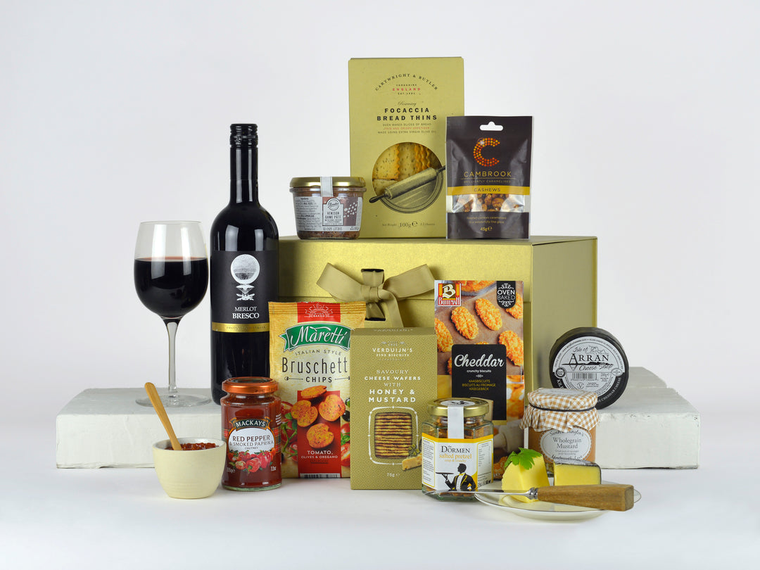 A bottle of red wine with savoury snacks such as bruschette bites, cheddar cheese bites, cashew nuts, chutney, pate, cheese and other savoury treats in a Gold Gift Box with Gold Ribbon