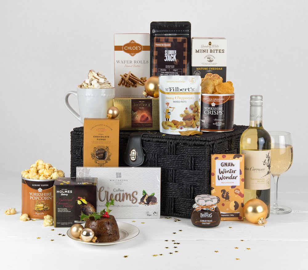 A bottle of white wine and christmas treats such as christmas pudding, christmas chutney, hot chocolate, biscuits, chocolates, popcorn and some savoury treats in a black gift basket