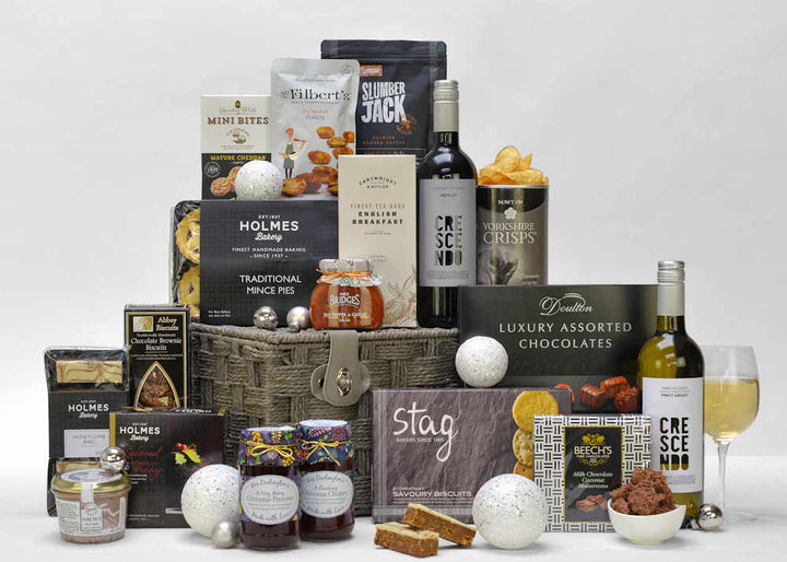 A bottle of red and white wine and christmas food items such as mince pies, christmas pudding, christmas preserve and chutney, tea, coffee, chocolates and savoury snacks in a grey gift basket