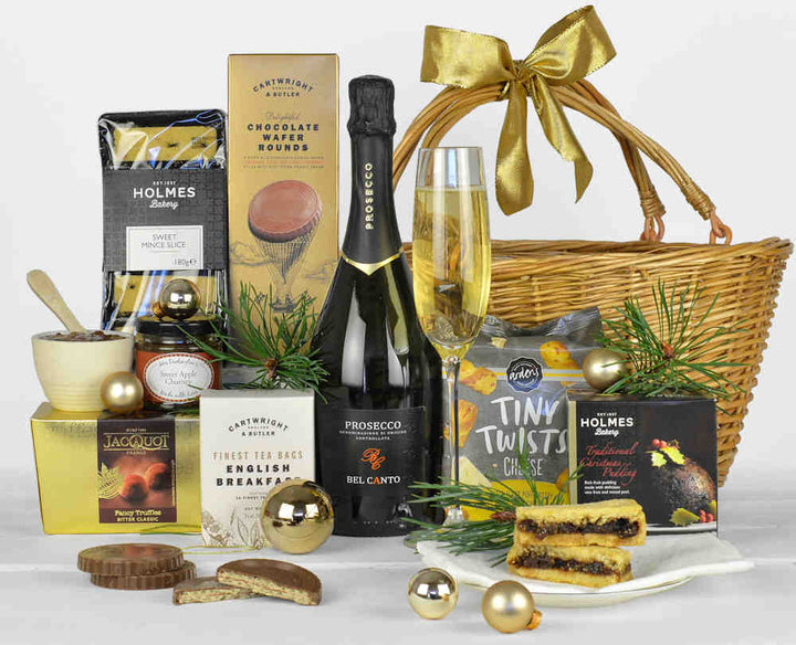 A bottle of prosecco with a christmas pudding, tasty savoury snacks, chocolates, tea, chocolate biscuits and sweet mince pie slices in a round wicker gift basket