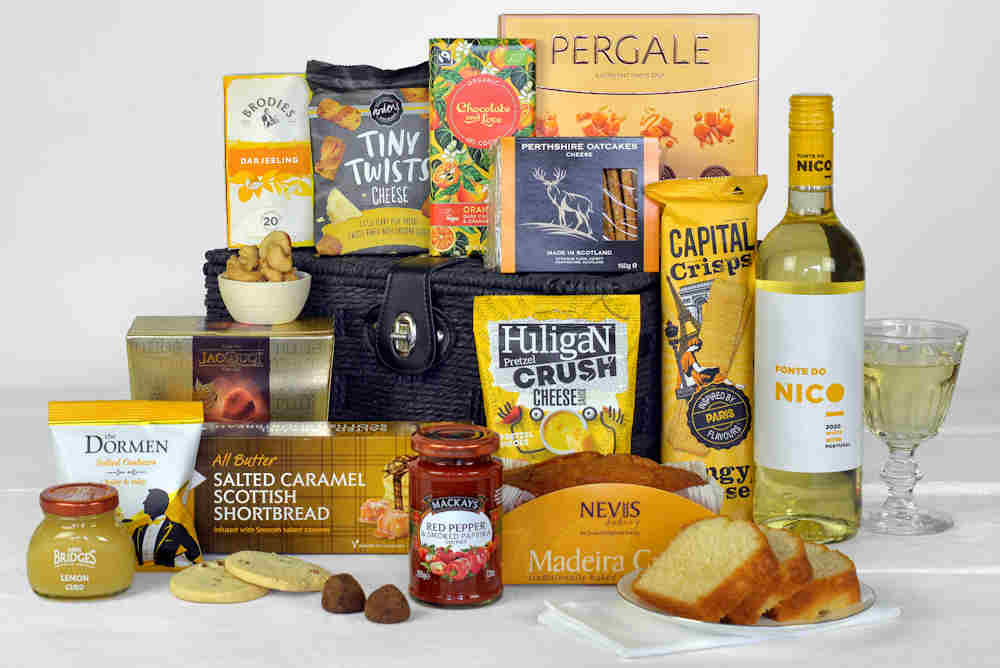 A bottle of white wine, savoury snacks, chocolates, biscuits, cake, preserves and a chutney in a black gift basket